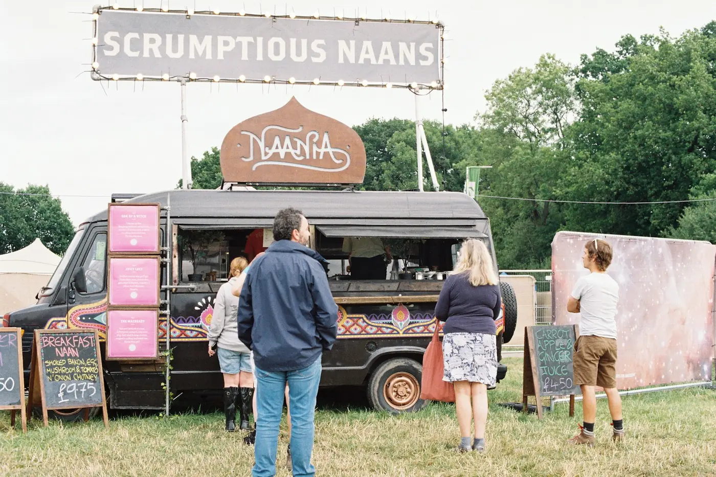 Black catering van at a festival with a big lettered header sign, pink menu panels and chalkboards, customers queuing at the hatch