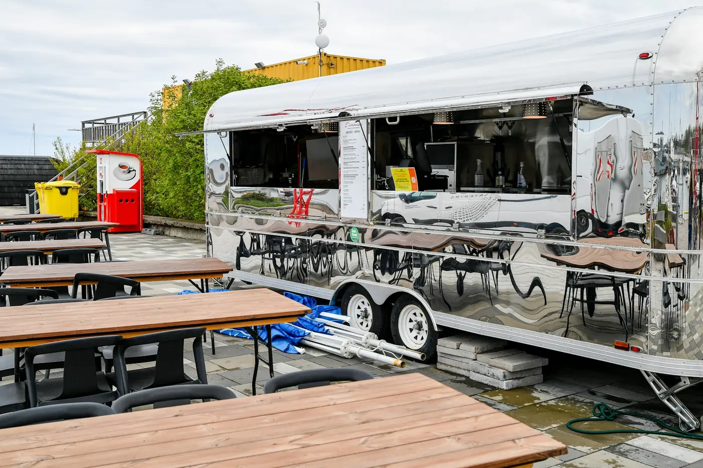 Polished stainless catering trailer with a long serving hatch and printed menu panels beside outdoor seating