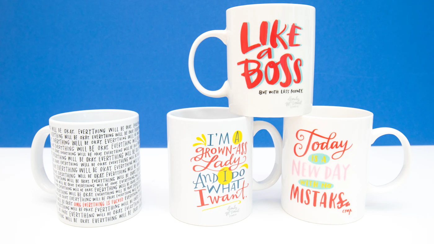 Four white ceramic mugs printed with black hand-lettered slogans on a blue background