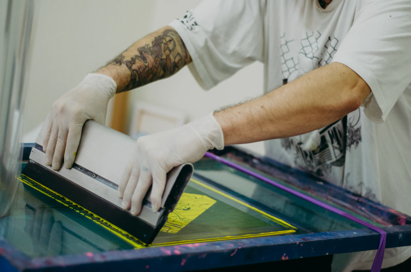Gloved hands pulling a squeegee across a silk screen during garment screen printing