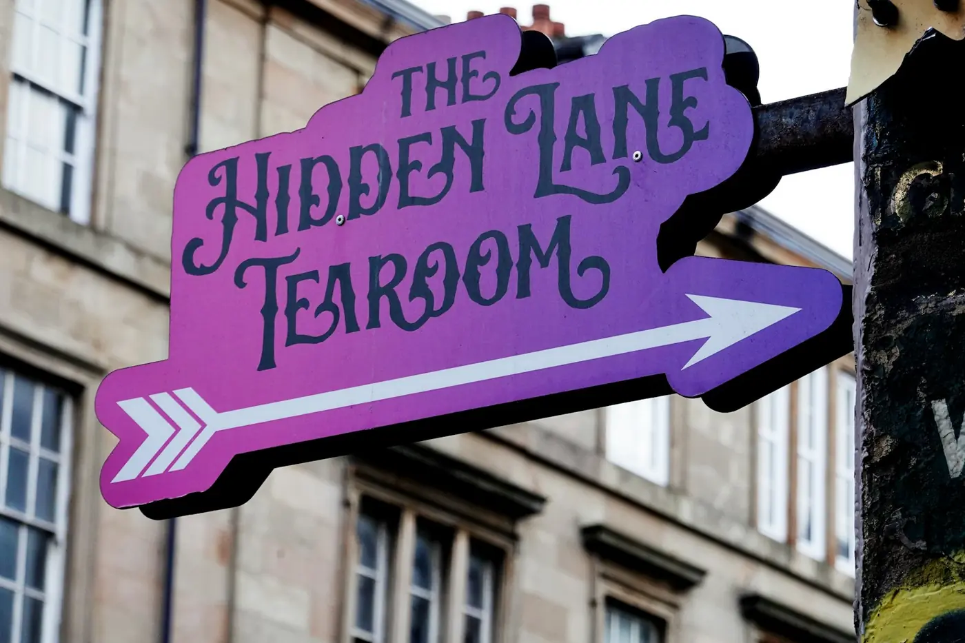 Purple shaped projecting sign reading The Hidden Lane Tearoom with an arrow, on a stone town centre building